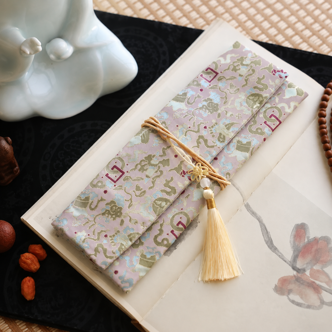 Blush pink silk incense carry pouch with green and pale blue auspicious brocade motifs, golden cord, and an ivory tassel with gold accent, placed on an open art book atop a dark cloth.