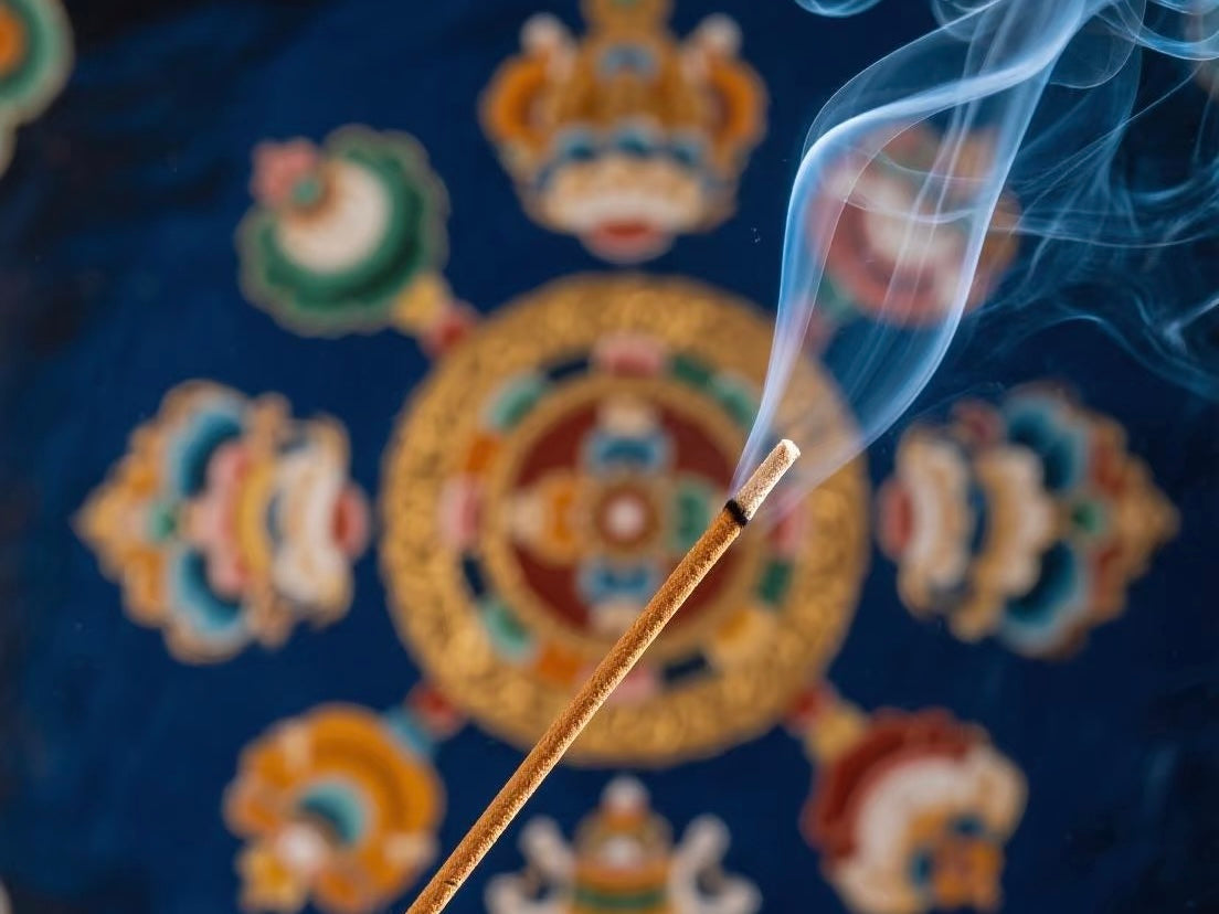 A Tibetan incense stick burning with delicate smoke rising in front of a colorful mandala design on a deep blue background.