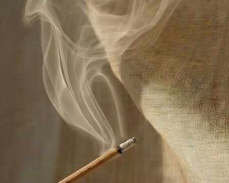A burning incense stick with a thin trail of smoke curling upwards against a textured, light beige background.