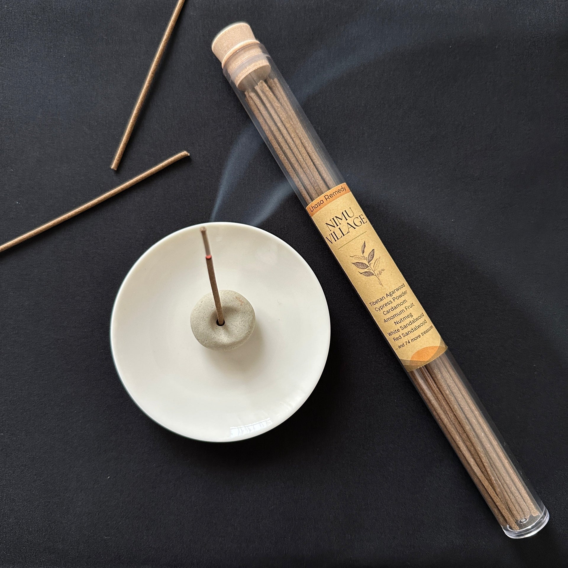 Nimu Village Tibetan incense sticks in a glass tube on a dark background with a white plate and a ceramic holder holding a burning incense stick.