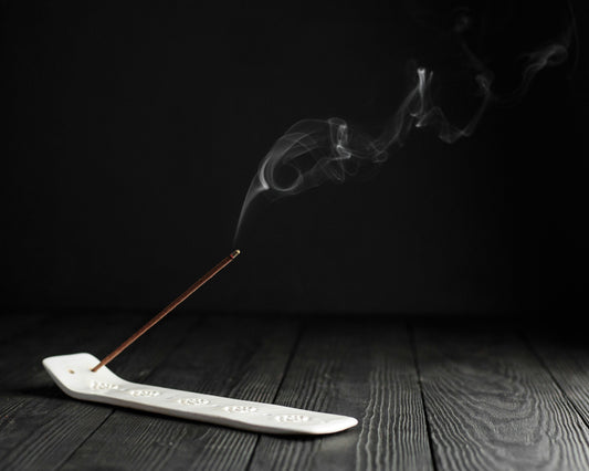 Burning incense stick placed on a white ceramic holder, with elegant smoke swirling into the air against a dark minimalist background, evoking a peaceful and meditative mood.