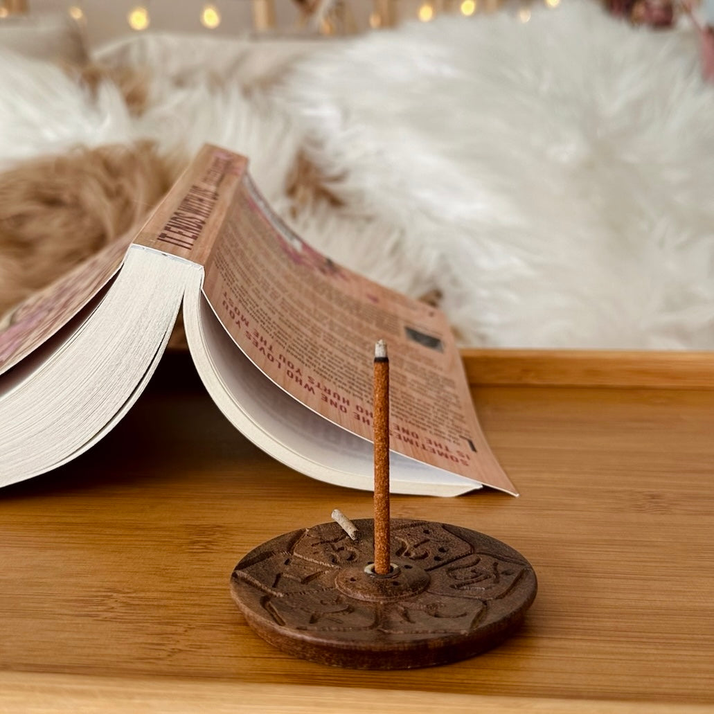 A burning Tibetan incense stick on a tray with a book. Pillows and lights are behind on a bed.