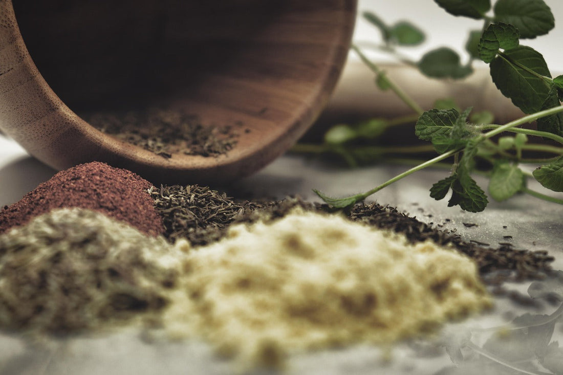 Close-up of natural herbal powders and dried leaves spilling from a wooden bowl, with fresh green sprigs beside them, symbolizing traditional Tibetan medicine and natural remedies.