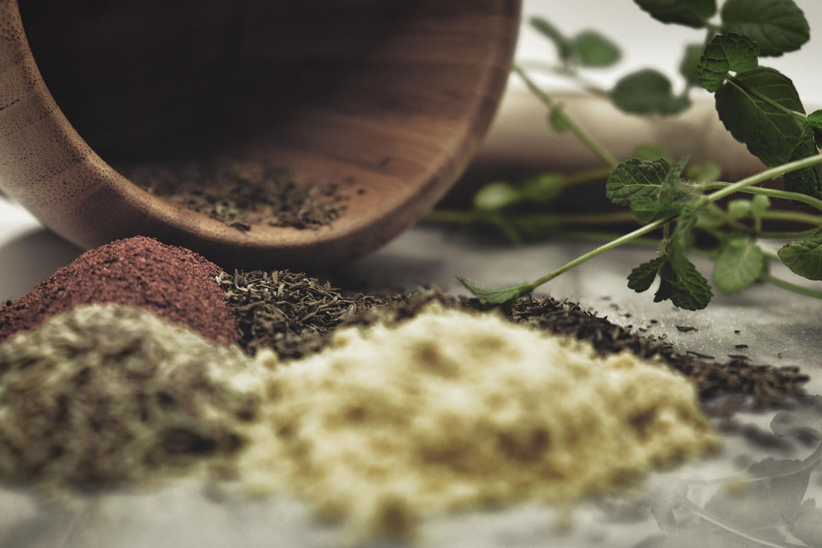 Close-up of natural herbal powders and dried leaves spilling from a wooden bowl, with fresh green sprigs beside them, symbolizing traditional Tibetan medicine and natural remedies.