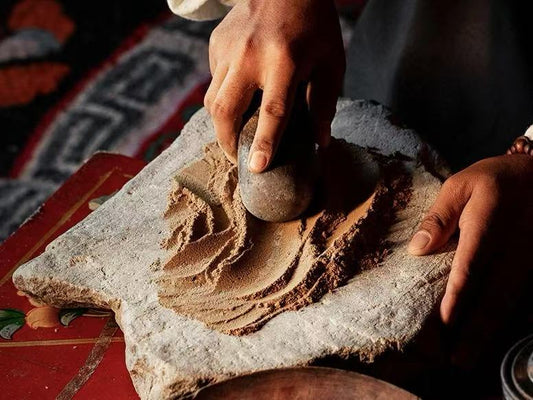 Close-up of hands grinding sacred Tibetan incense herbs using a traditional stone mill, capturing the ancient artisan process.