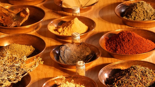 A variety of natural Tibetan incense ingredients, including ground herbs, woods, and resins, neatly organized in traditional copper bowls on a wooden surface.
