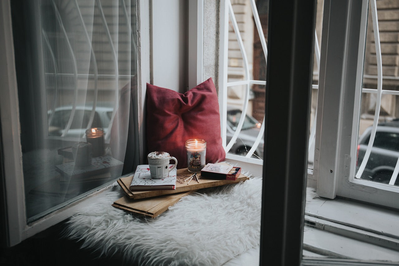 A cozy window nook featuring a lit candle, books, and a warm drink, illustrating why some people prefer candles over incense for relaxation rituals. Highlights the candles vs incense preference in spiritual and daily routines.