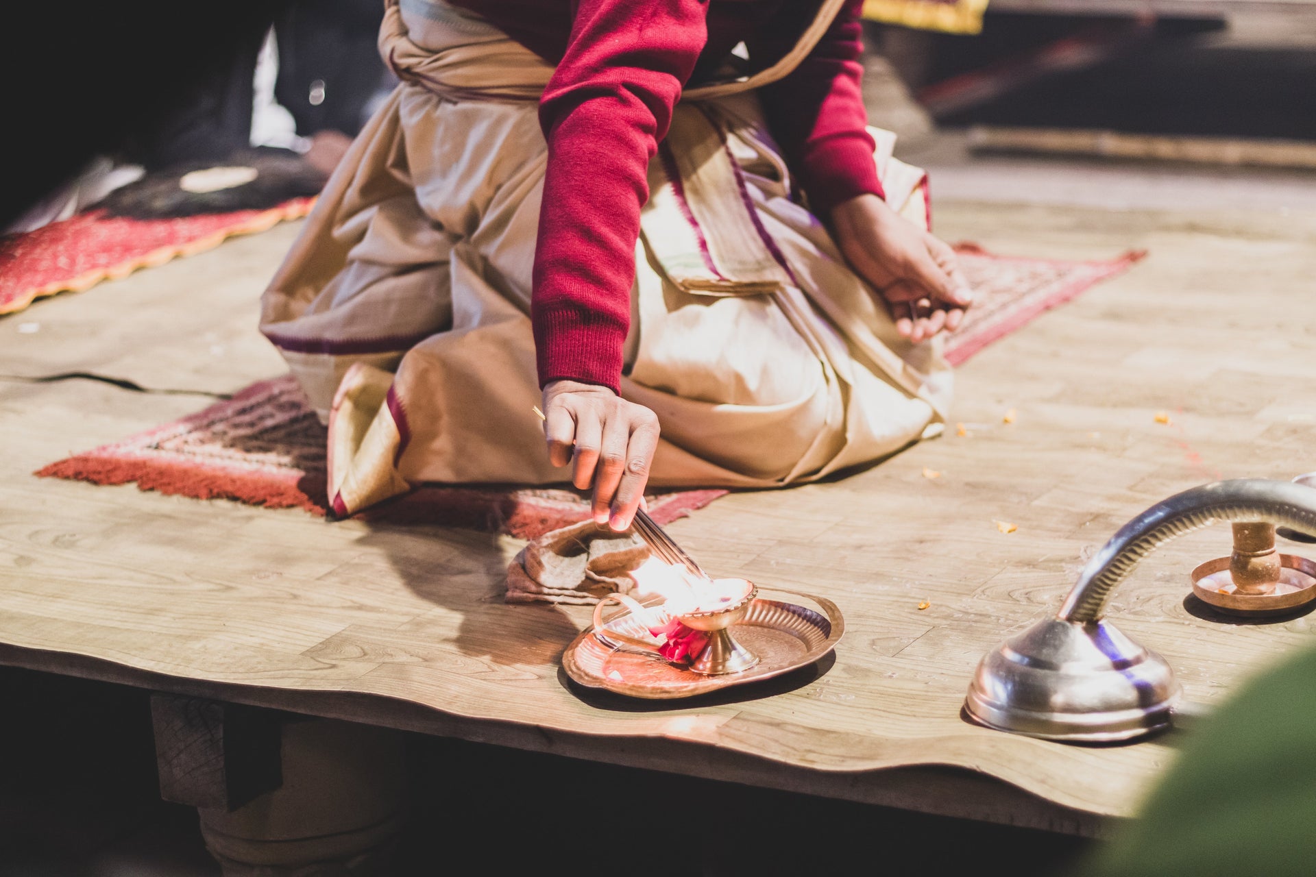 Close-up of a woman in traditional attire lighting incense sticks as part of a ritual, with a copper plate and ceremonial setup on a wooden floor.