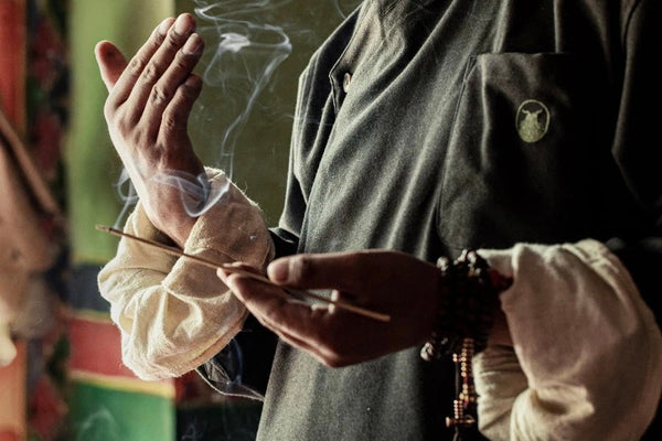 A man stands calmly, holding a burning Tibetan stick emitting wisps of smoke, embodying peace and spiritual reflection in a tranquil setting.