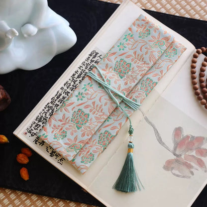 Light jade silk incense carry pouch with coral lotus vine brocade pattern and a soft green tassel, placed on an open art book atop a dark tray