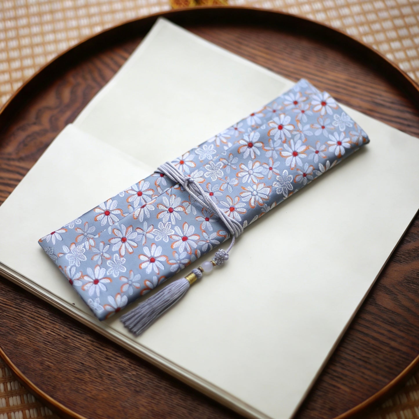 Soft grey silk incense carry pouch with daisy floral brocade pattern and a pale grey tassel, placed on an open art book atop a wooden tray
