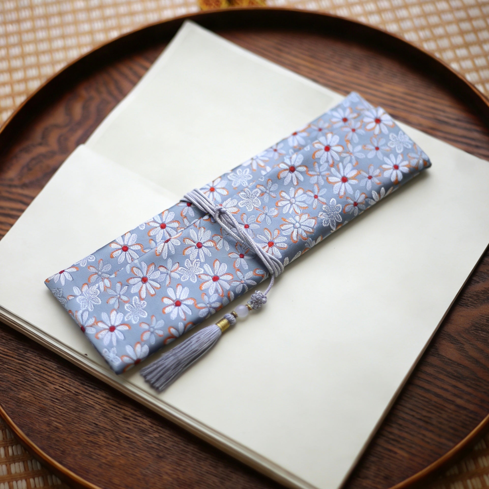 Soft grey silk incense carry pouch with daisy floral brocade pattern and a pale grey tassel, placed on an open art book atop a wooden tray