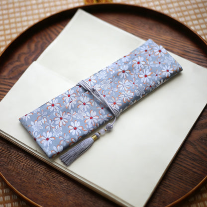 Soft grey silk incense carry pouch with daisy floral brocade pattern and a pale grey tassel, placed on an open art book atop a wooden tray