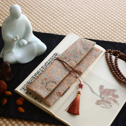 Sand-colored silk incense carry pouch with ruyi court brocade pattern and a warm coral tassel, placed on an open art book atop a dark cloth