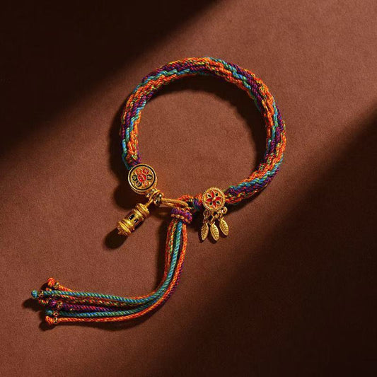Hand-braided Tibetan five-color blessing bracelet with a golden mantra prayer wheel charm and Dharma wheel pendants, displayed on a warm brown background.