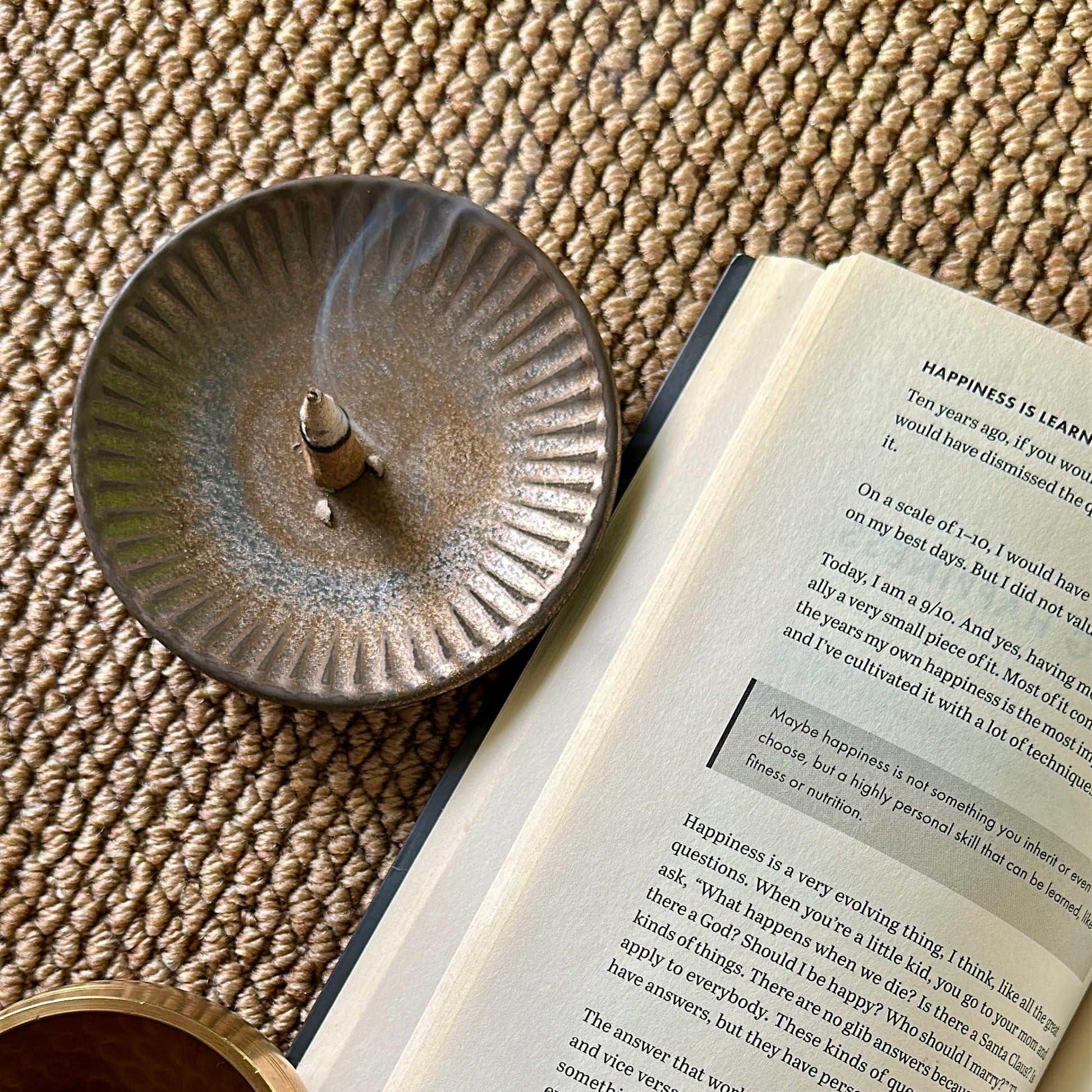 Sera Serene Tibetan incense cone gently burning in a ceramic holder beside an open book about happiness, creating a peaceful reading ritual setup on woven mat.