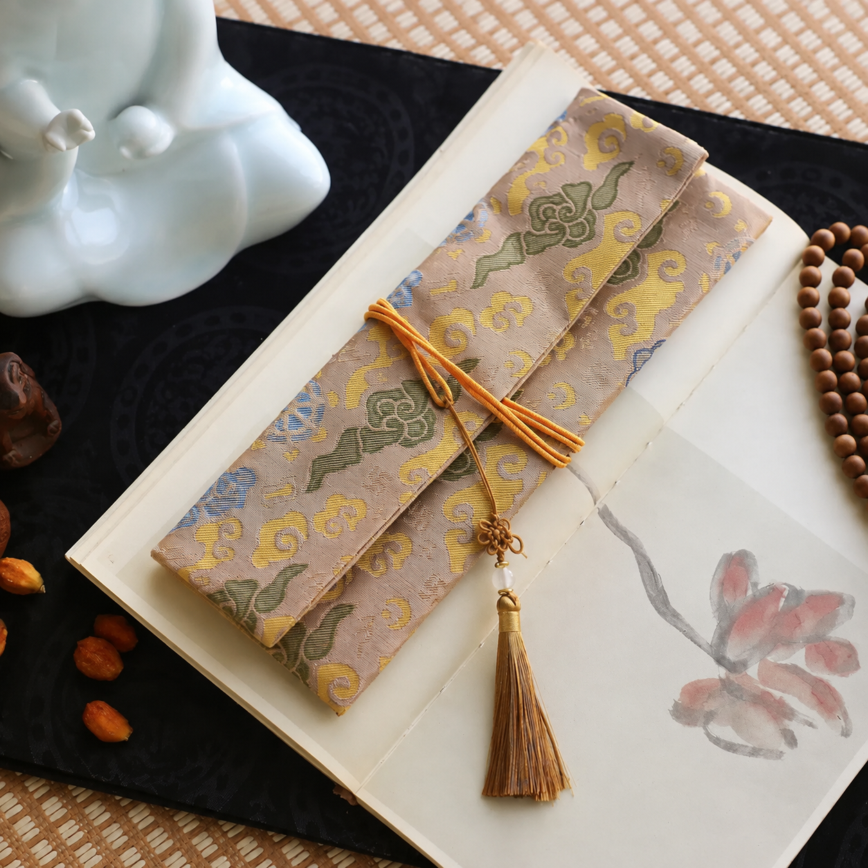 Taupe silk incense carry pouch with golden ruyi cloud brocade pattern, orange cord, and a golden brown tassel, placed on an open art book atop a dark cloth