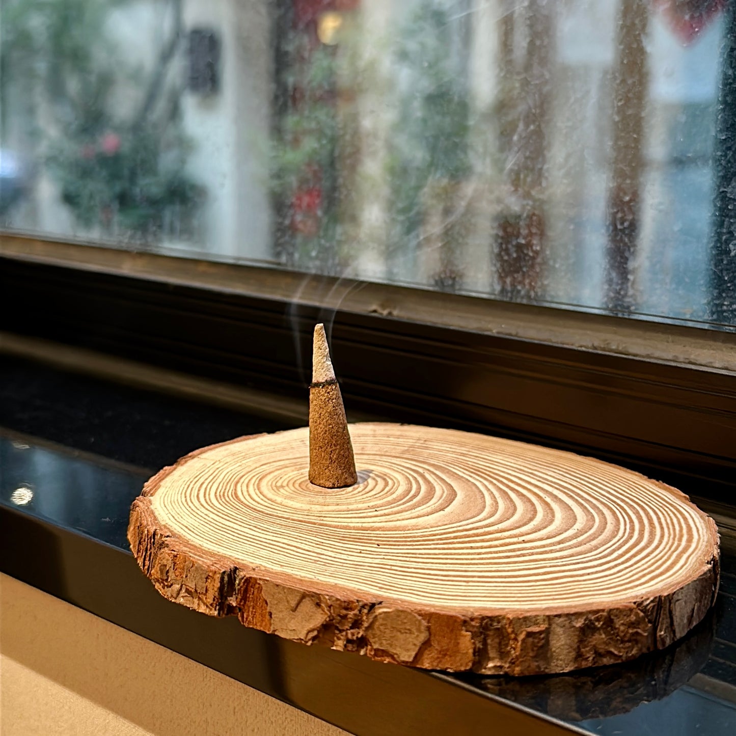 Lucky Zaki Tibetan incense cone gently burning on a natural wood slice holder by the window, releasing a thin trail of smoke.