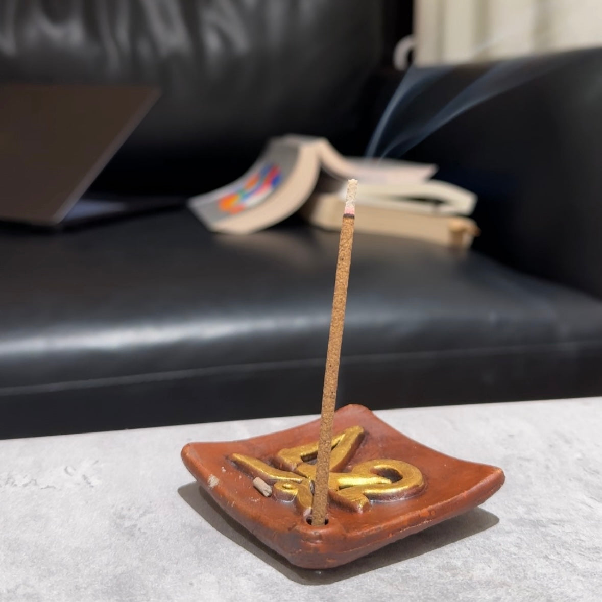 Potala Palace Tibetan incense stick gently burning in the Sacred OM Ceramic Incense Holder, with soft smoke rising and an open book resting on a black leather sofa in the background.