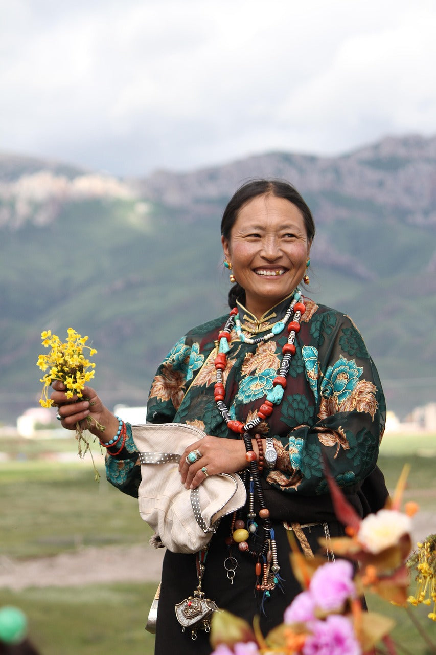 Smiling Tibetan woman in traditional clothing holding wildflowers in a highland valley landscape