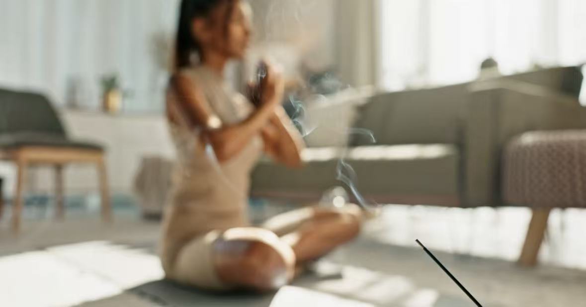 A blurred woman meditating in the background with focus on a smoking Tibetan incense stick in front